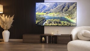 Una sala de estar moderna con un televisor de pantalla plana mostrando un paisaje montañoso, un jarrón blanco con plumas de pampa, un sillón blanco y mullido, y un mueble de madera oscura con objetos decorativos.