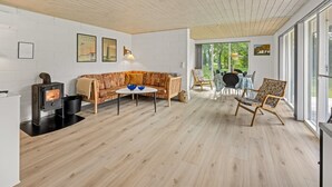 Ein modernes Wohnzimmer mit Holzboden, einer Couch, einem Couchtisch und einem Essbereich mit Stühlen und Tisch. Ein Kamin und ein großes Fenster bieten einen Blick ins Freie.