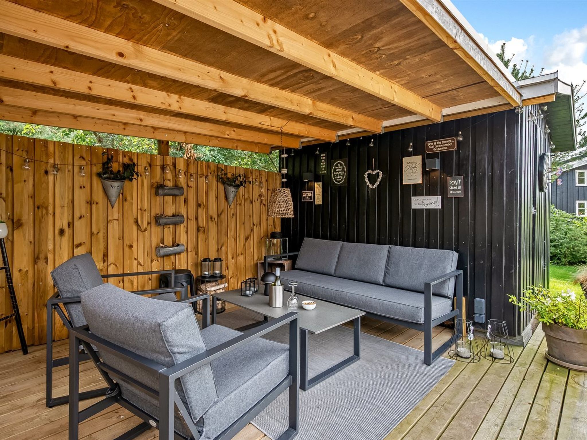 An outdoor seating area with a wooden deck, a black wall with various signs, and a grey sofa.