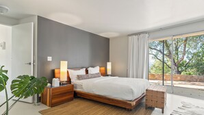 Una camera da letto moderna con un letto grande, comodini, un tavolino da notte, una pianta in vaso e vista sull'esterno tramite una porta scorrevole in vetro.