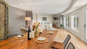 Un amplio comedor con una mesa de madera dispuesta para cuatro personas, rodeada de sillas. Hay velas y una botella de vino sobre la mesa, y un jarrón con flores. La habitación tiene ventanas grandes y vistas al exterior.