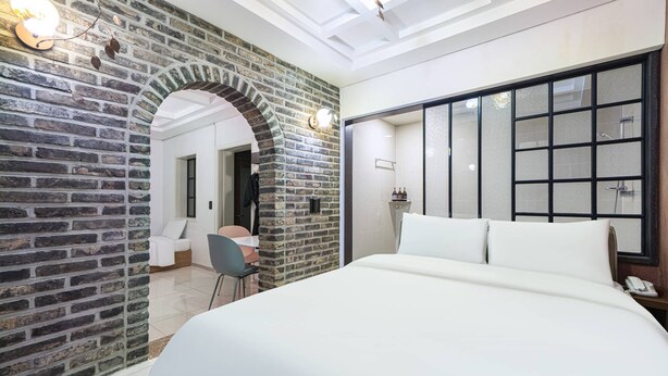 A modern hotel room with a brick wall, a large bed, a desk with a chair, and a large window.