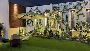 A well-lit outdoor patio area with artificial grass, a seating arrangement, and a variety of potted plants.
