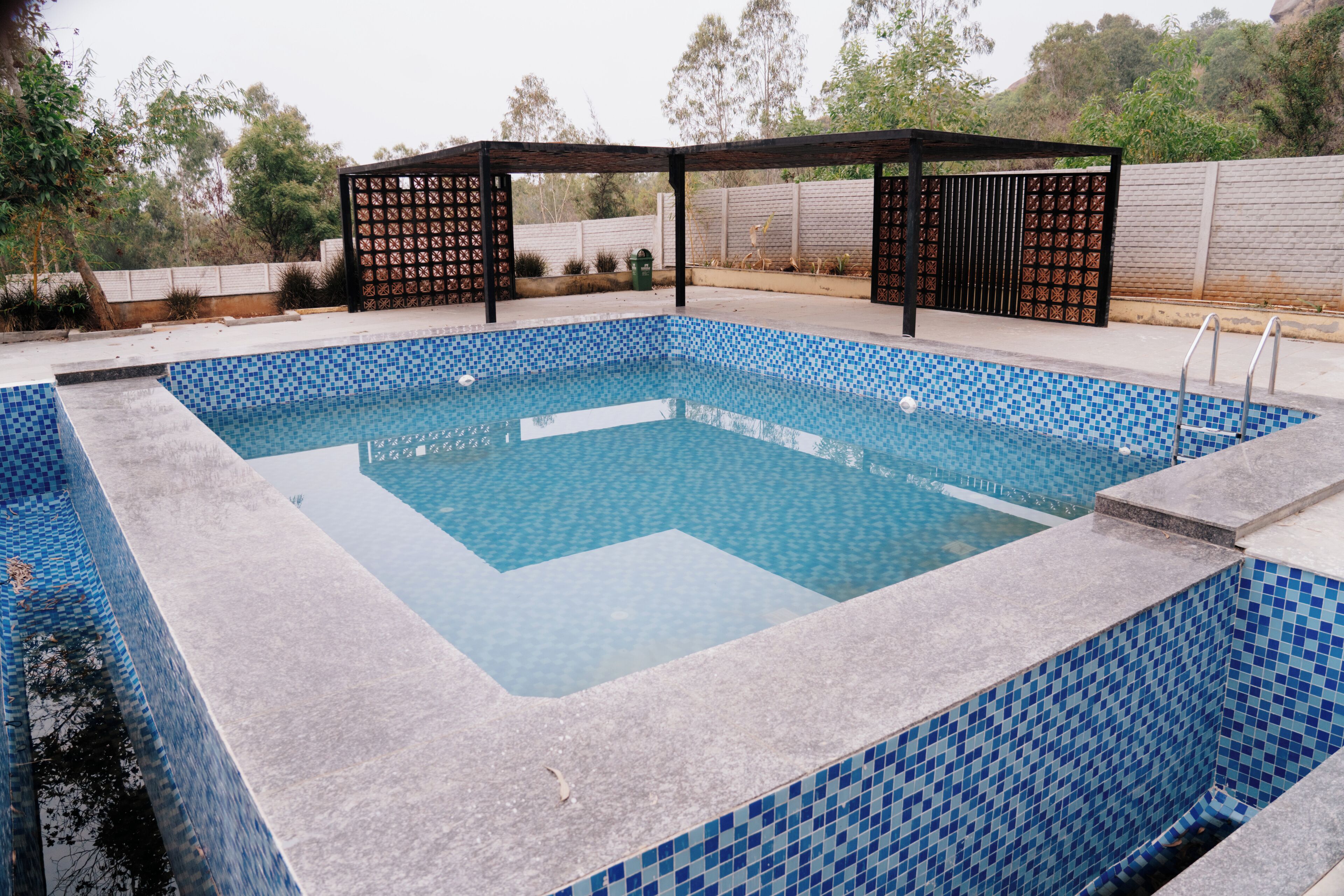 Une piscine rectangulaire aux carreaux de mosaïque bleus, entourée d’une terrasse en pierre et d’une pergola en bois.