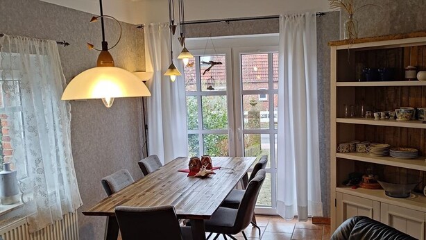 Een eethoek met een houten tafel, stoelen en hanglampen. Er is een groot raam met gordijnen en uitzicht op groen buiten. Een houten boekenkast met servies en decoratieve objecten is zichtbaar.