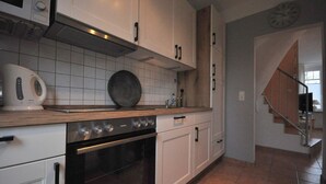 A kitchen with white cabinets, a stove, an oven, and a microwave. There is a clock on the wall and a staircase leading to another room. - Land-Domizil terraced house 1 - Terraced house Land-Domizil 1 (Werdum)