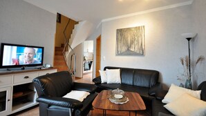 Ett modernt vardagsrum med en svart lädersoffa, ett träbord och en tv-skärm som visar en person. En trappa leder upp till ett kök som syns i bakgrunden.