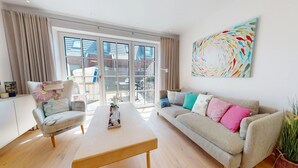 Een moderne woonkamer met een bank, een salontafel en een stoel. Er is een groot raam met gordijnen, en een kleurrijk abstract schilderij aan de muur.