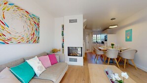 Een moderne woonkamer met een bank, een eethoek met stoelen, een open haard en een keuken op de achtergrond.