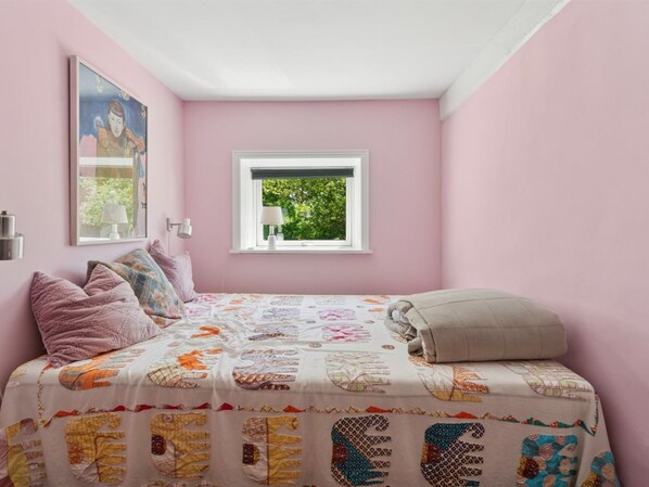 A bedroom with a pink wall, a window with a view of greenery, a bed with a colorful quilt, and a framed picture on the wall.