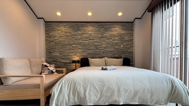 Un dormitorio moderno con una pared de acento de piedra, una cama con colcha color beige, una silla de madera y una mesita de noche con lámpara.