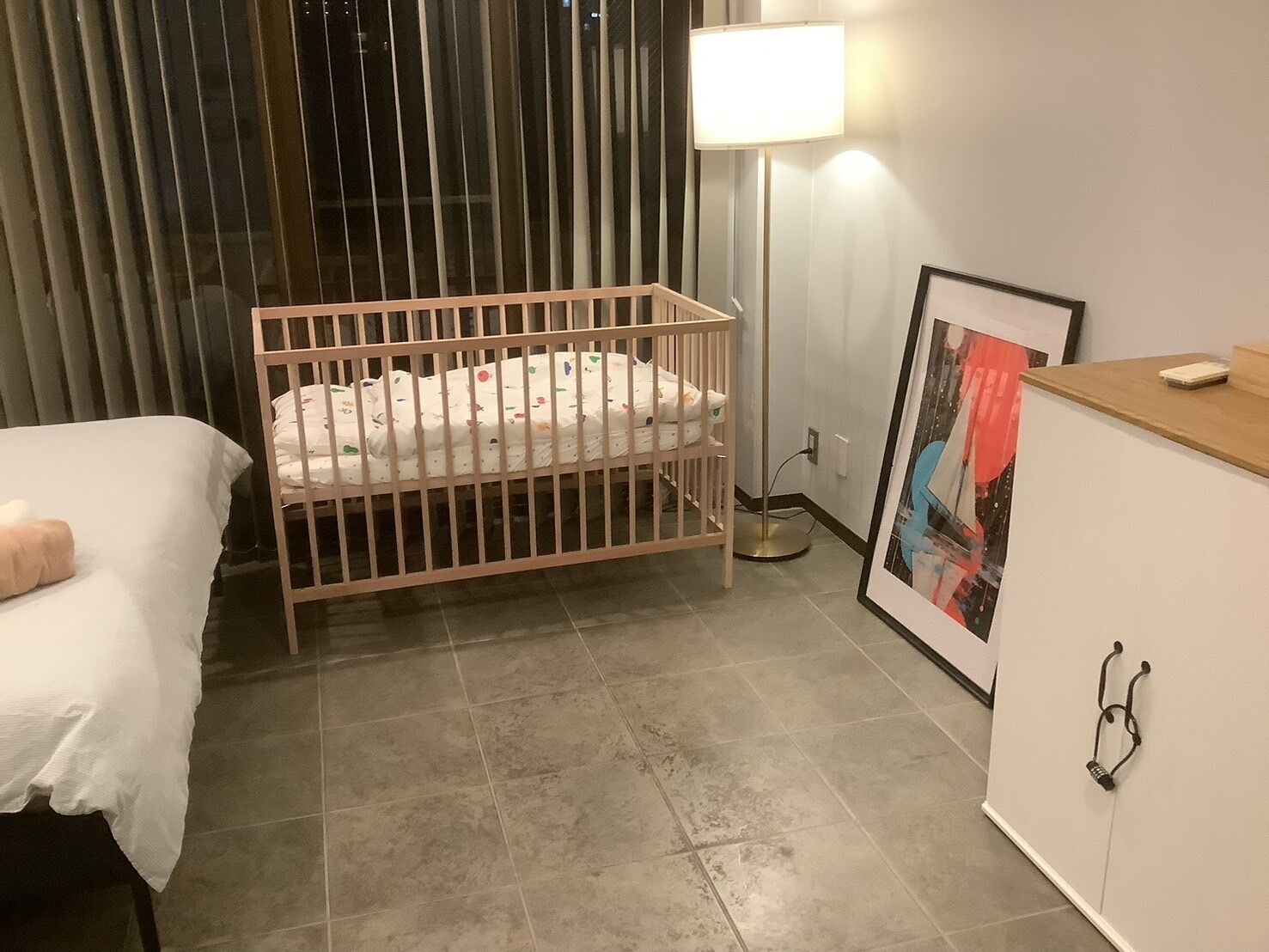 A room with a crib, a bedside table, a lamp, and a framed picture on the wall.