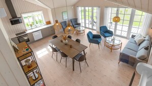 A spacious living area with a dining table, blue armchairs, and a sofa. There are large windows providing natural light and a view of the outdoors.