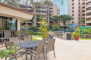 An outdoor patio with a round table and chairs, surrounded by potted plants and a barbecue grill. - Valley Isle Resort #104 - Vacation West Maui (Lahaina)