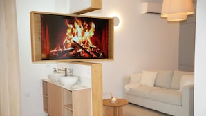 A modern bathroom with a wooden vanity, a white sink, and a flat-screen TV mounted above the vanity displaying a fireplace.