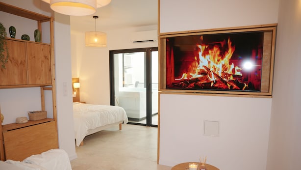 A modern hotel room with a bed, a sofa, a small table with candles, a wooden shelf with decorative items, and a large TV screen displaying a fire.