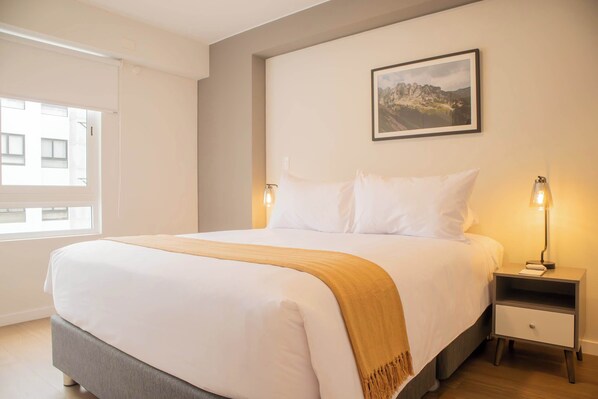 A neatly made bed with a white comforter and a beige throw blanket, a bedside table with a lamp, and a framed picture on the wall.