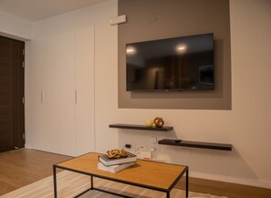 A modern living room with a flat-screen TV mounted on the wall, a wooden coffee table with books and a pretzel, and a small shelf with decorative items.