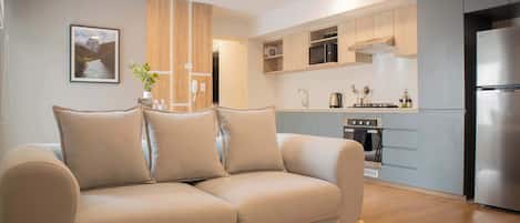 A modern living room with a beige sofa, a coffee table with a book and a pretzel, and a kitchen area with light blue cabinets and stainless steel appliances.