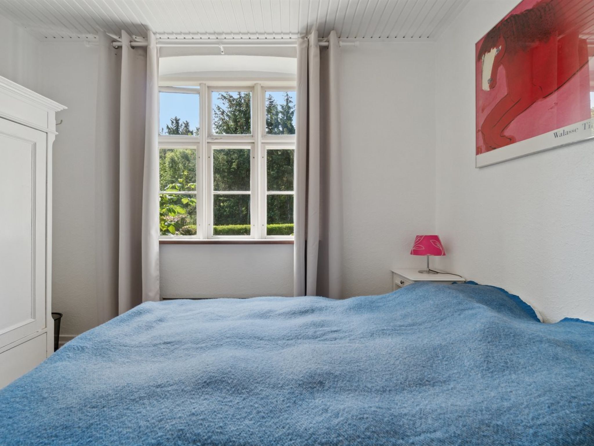 Une chambre avec une grande fenêtre, un lit recouvert d’une couverture bleue, une table de chevet avec une lampe et une affiche accrochée au mur.