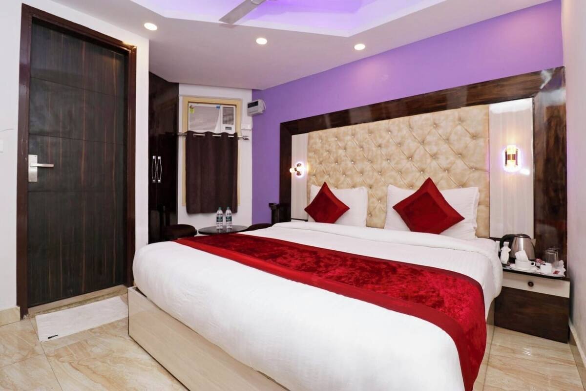 Habitación de hotel con una cama grande, cabecera de madera, manta roja y mesita de noche con lámpara.