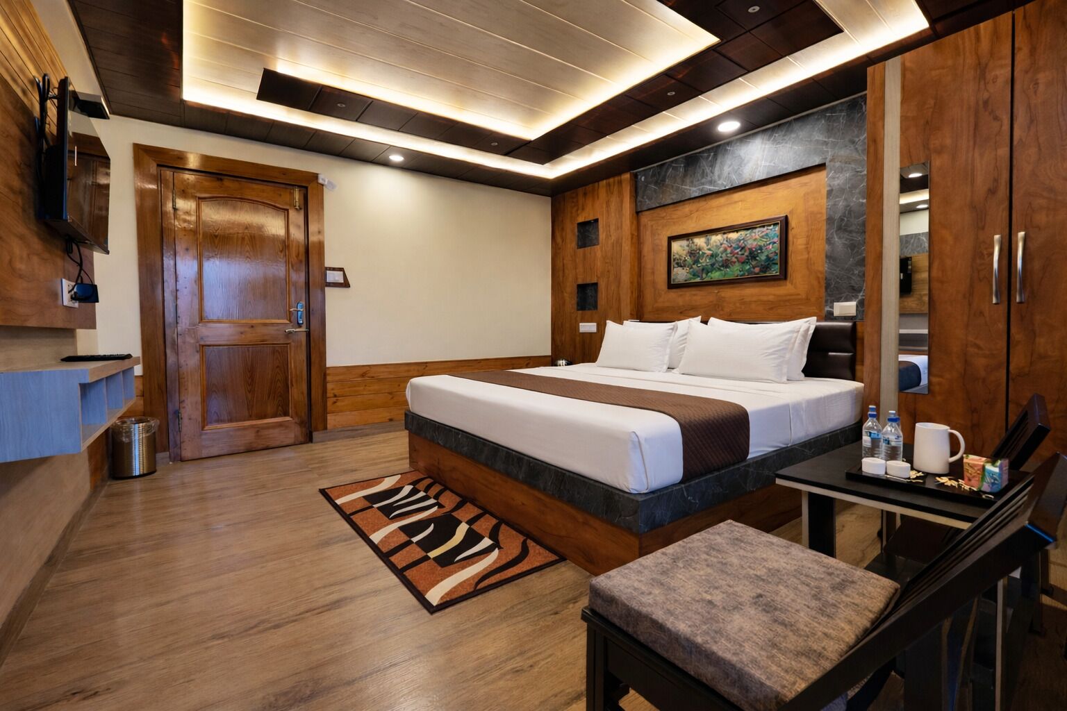 A hotel room with a wooden floor, a bed with white bedding, a desk with a chair, a small trash bin, and a painting on the wall.