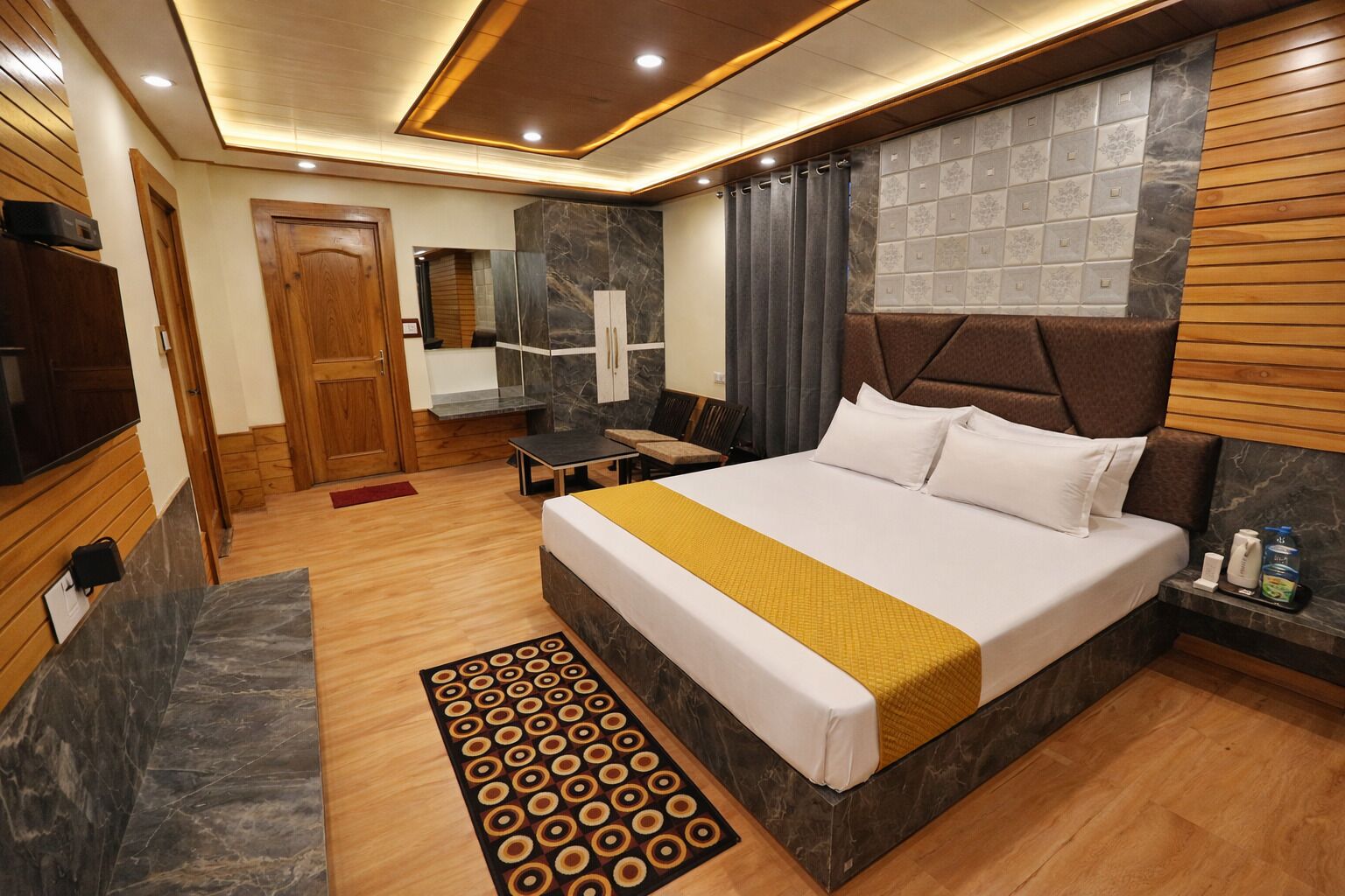 A hotel room with a large bed, wooden headboard, marble wall, and a small table with a bottle.