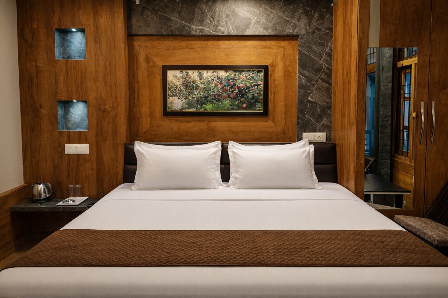 A hotel room with a large bed, a painting above the headboard, a wooden wardrobe, and a nightstand with a kettle.