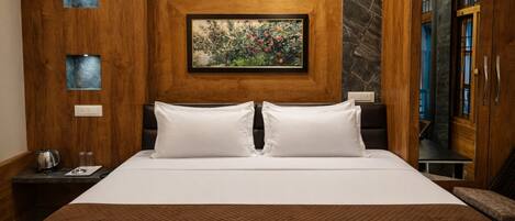 A hotel room with a large bed, a painting above the headboard, a wooden wardrobe, and a nightstand with a kettle.