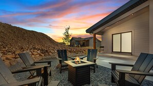 An outdoor seating area with Adirondack chairs and a fire pit, set against a backdrop of a sunset and a stone wall.