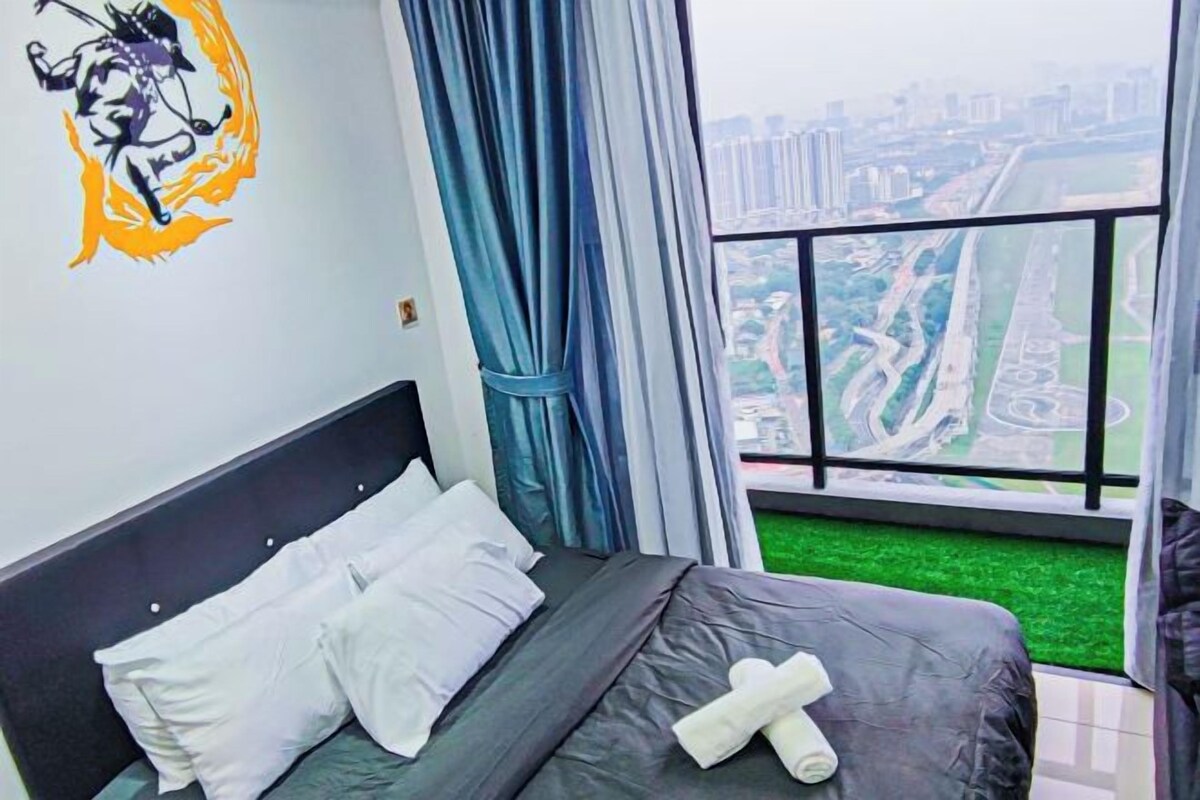 Un dormitorio con cama, almohadas blancas, una toalla, una ventana que ofrece vistas a un paisaje urbano y césped artificial verde.