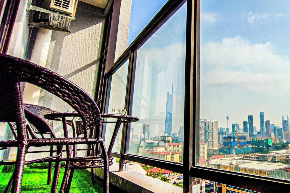 Ein Balkon mit Korbstühlen, einem Glastisch und Blick auf die Stadt durch große Fenster.