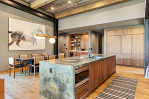 A modern kitchen with a marble island, wooden cabinetry, and a dining area with chairs.