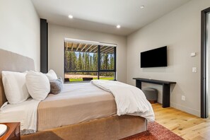 A modern bedroom with a large bed, a TV mounted on the wall, and a view of a forest through a large window. - Ski-In/Ski-Out | In-Ground Hot Tub (Big Sky)