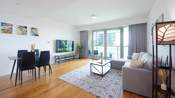 A modern living room with a sofa, a coffee table, a TV, and a dining area with chairs and a table. - Silk Valley - Luxury 1B near Raha Beach (Abu Dhabi)