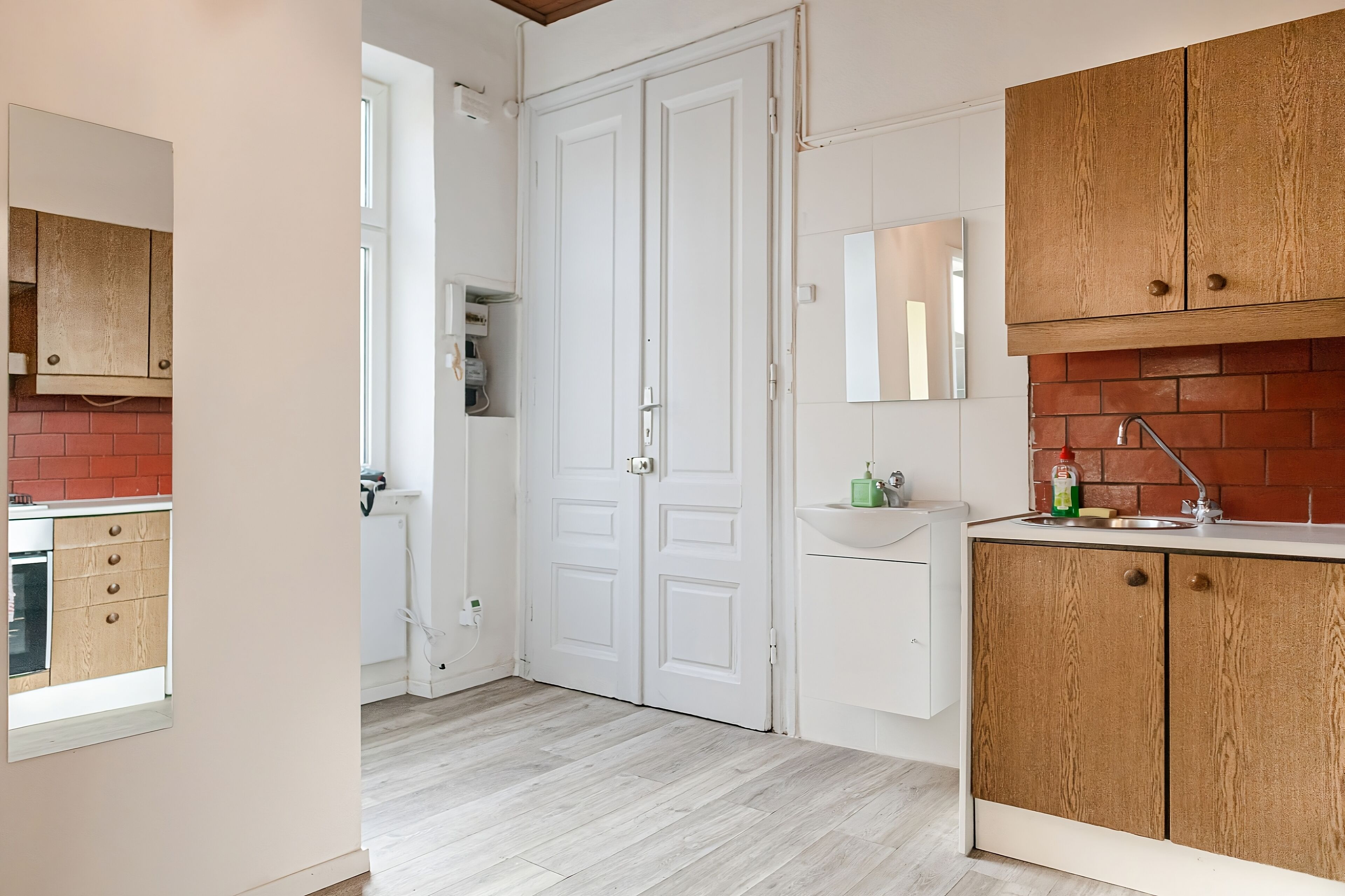 Een keuken met witte kasten, een tegelwand van baksteen, en een dubbele deur die naar een andere kamer leidt.