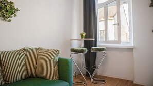 Ein grünes Sofa mit gestreiften Kissen, zwei Metall-Barhocker mit Sitzpolstern, ein kleiner Tisch mit einer Pflanze und ein Fenster mit Blick auf Gebäude.