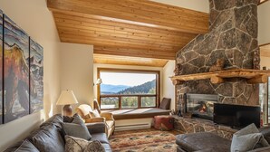 A cozy living room with a stone fireplace, leather sofas, and a large window offering a mountain view. - Walk to Explorer Gondola | Private Sauna (Big Sky)