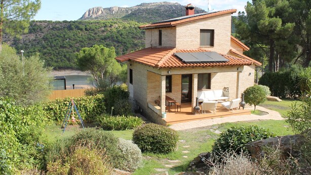 A two-story house with a patio area, surrounded by greenery and a mountain in the background.
