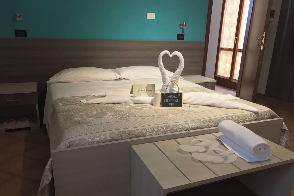 A neatly made bed with a heart-shaped towel, a sign reading "Chambre," and a rolled towel on a bedside table.