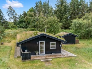 Eine kleine, dunkle Holzhütte mit geneigtem Dach, einer Tür und einem Fenster. Vor der Hütte befindet sich eine kleine Veranda mit Stufen, die zur Tür führen. Die Hütte ist von einer Rasenfläche mit Bäumen im Hintergrund umgeben.