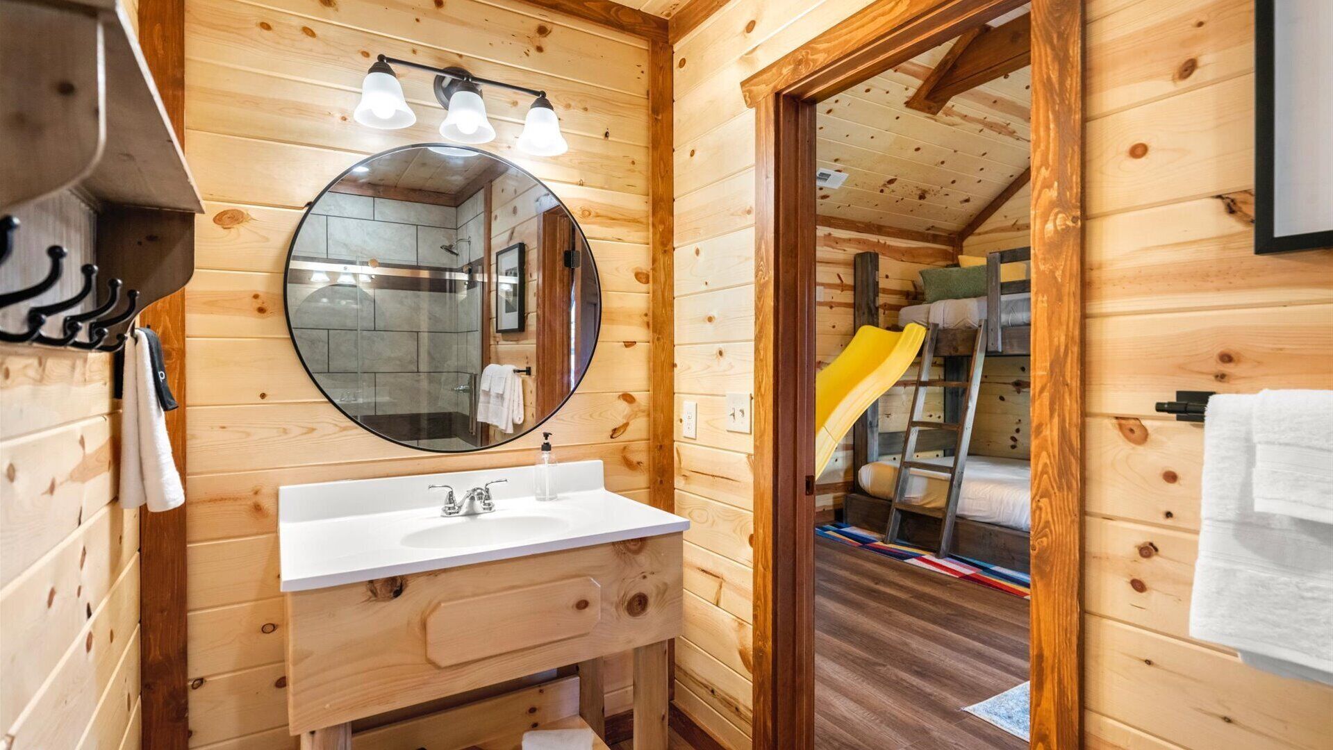 Interior de una cabaña de madera con un baño que incluye un espejo redondo, un lavamanos blanco y una zona de ducha.