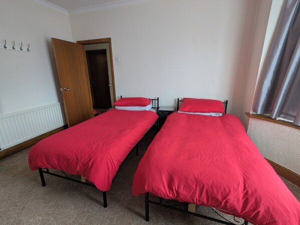 Una habitación con dos camas individuales, cada una con una colcha roja y almohadas a juego, una puerta de madera, un radiador y una ventana con cortinas.