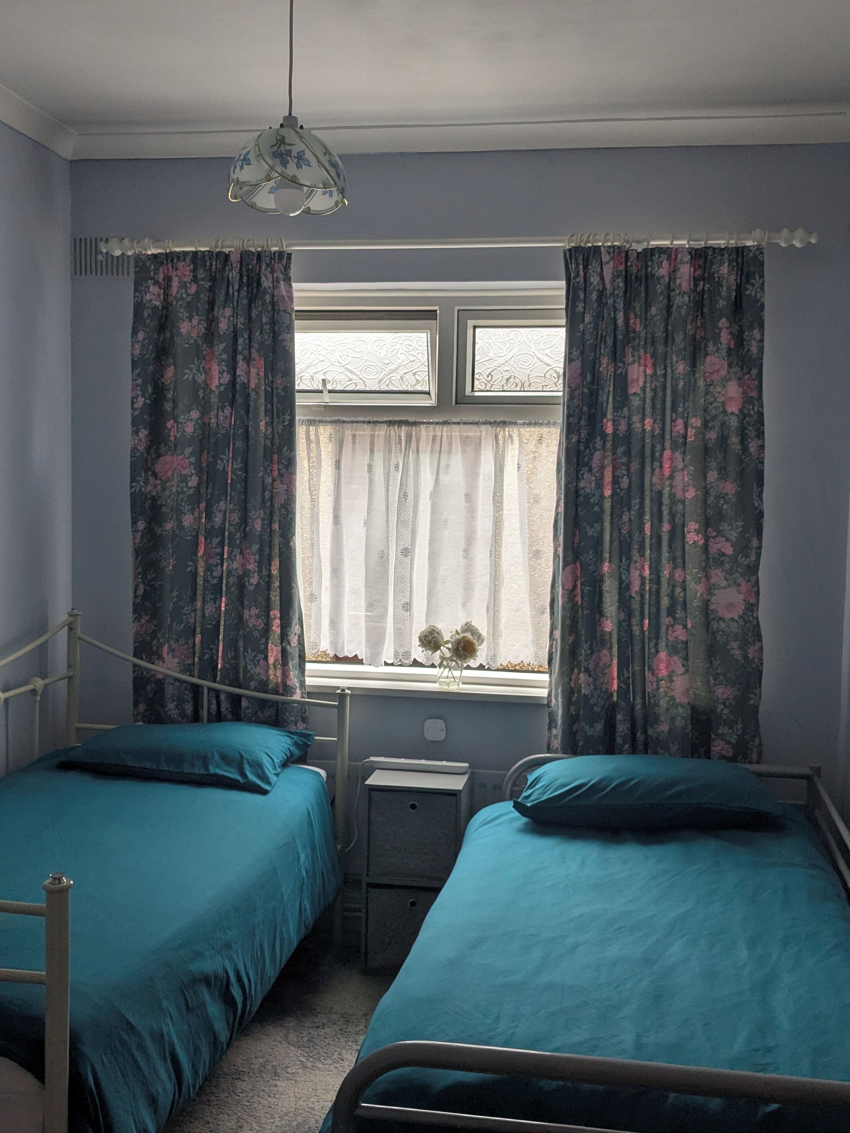 A bedroom with two single beds, a window with floral curtains, a bedside table, and a hanging light fixture.