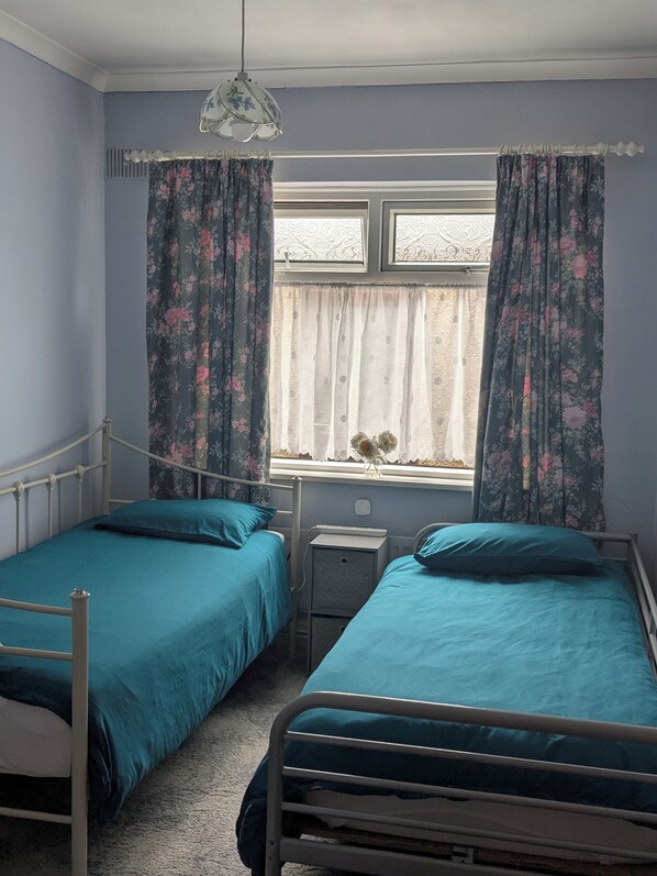 Un dormitorio con dos camas individuales, una mesita de noche y un ventanal con cortinas florales.