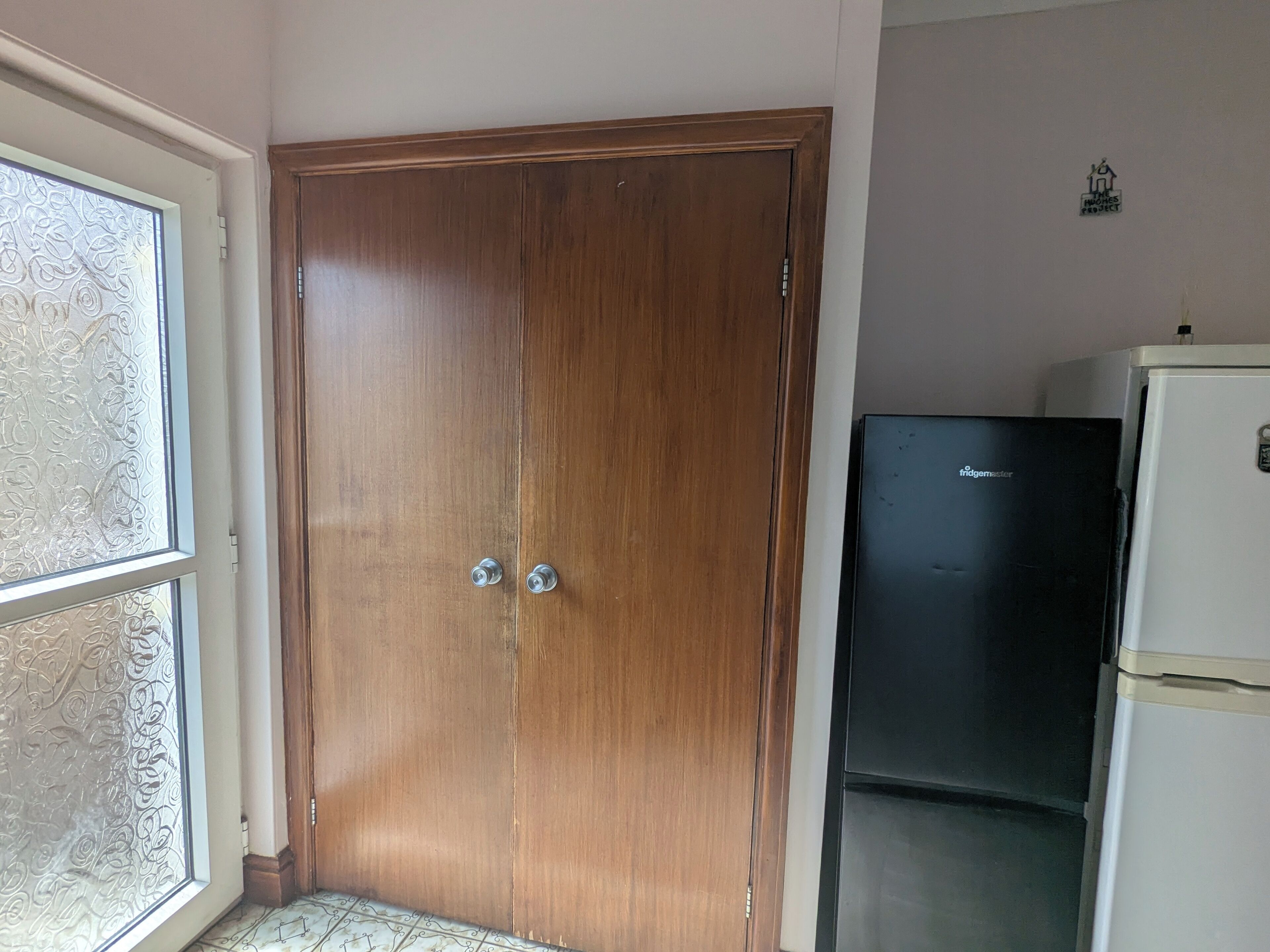 Ein Zimmer mit einem Holzschrank, einem schwarzen Kühlschrank und einem Fenster mit dekorierter Scheibe.