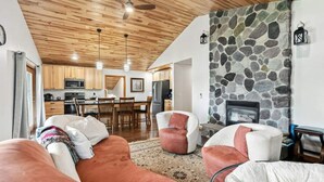 A cozy living room with a stone fireplace, wooden ceiling, and a dining area with a kitchen in the background. - Lunar Lodge on Windfall Lake (Exeland)