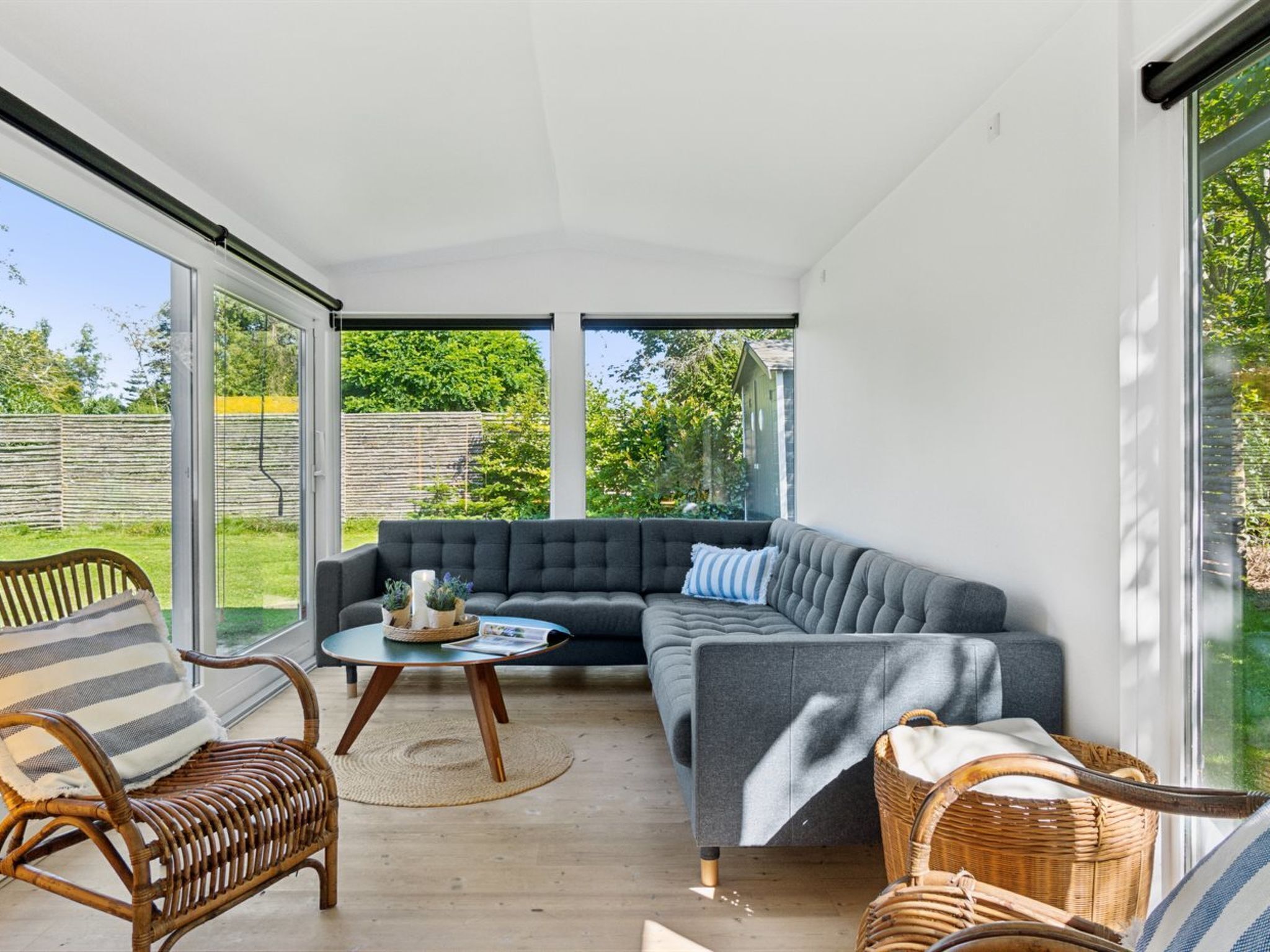 Ein modernes Wohnzimmer mit einem grauen Ecksofa, einem Korbstuhl, einem kleinen runden Tisch und einer Glastür, die zu einem Garten führt.