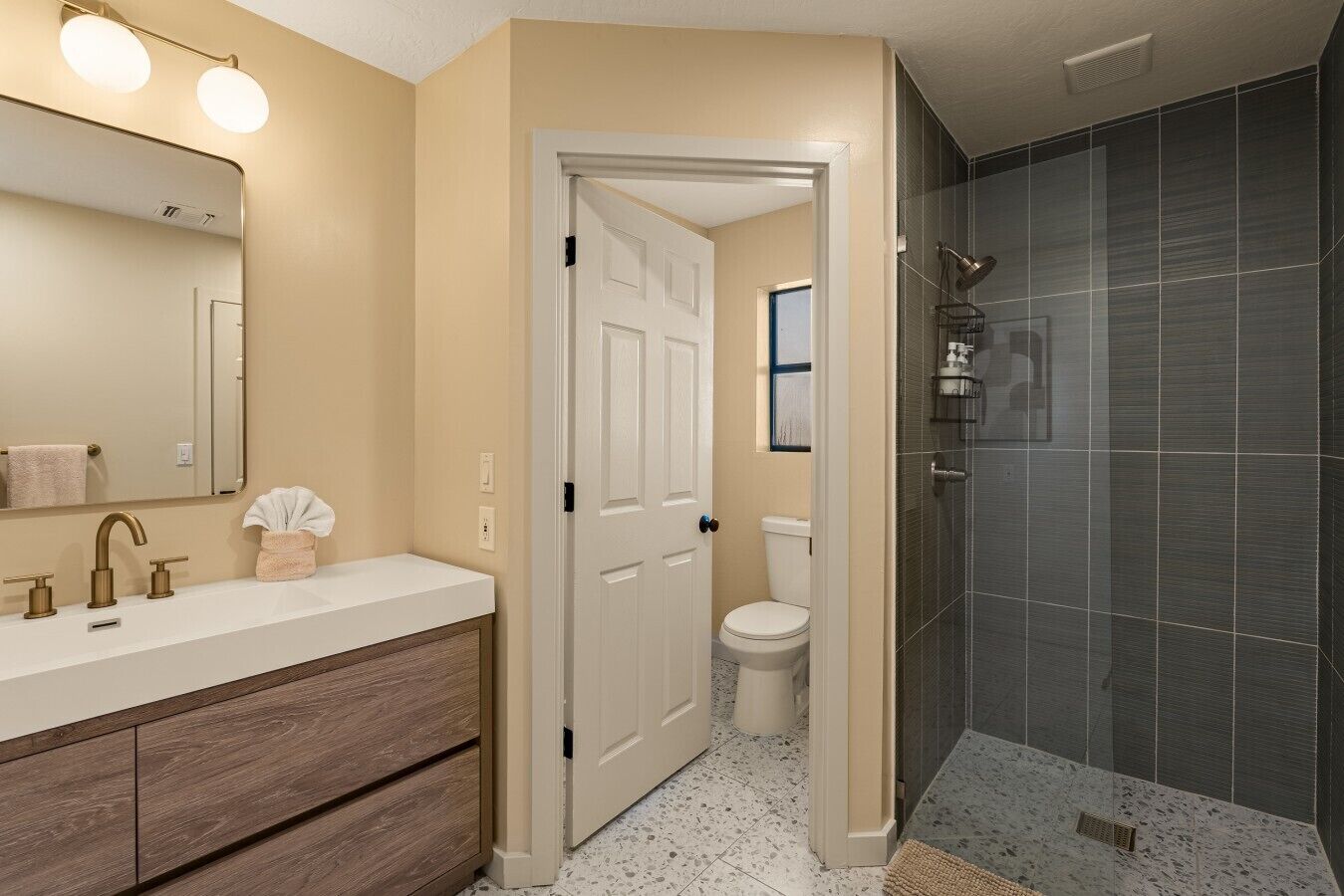 A bathroom with a white vanity, a mirror with lights, a shower with dark tiles, and a door leading to another room.