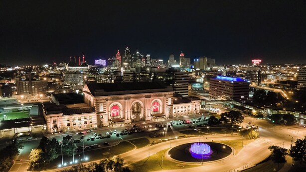 A cityscape at night with a prominent building featuring arched windows and a central fountain.